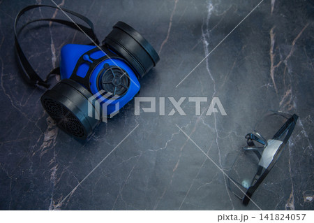 A blue respirator and goggles are spaced apart on a dark marble table. Concept, minimalism in labor protection, respiratory and visual protection, free space for text, safety. High quality photo A blue respirator and goggles are spaced apart on a dark marble table. Concept, minimalism in labor protection, respiratory and visual protection, free space for text, safety. High quality photo 141824057