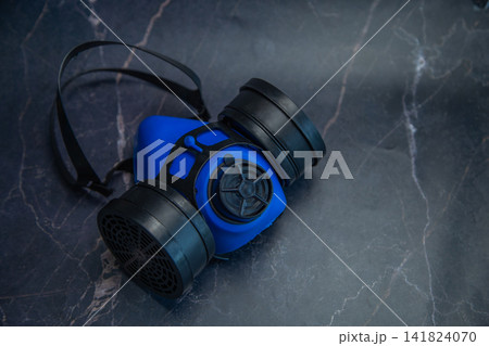 A blue half-mask respirator with black filters and elastic straps lies diagonally on a dark marble background. Concept of respiratory protection, industrial safety, reliable equipment. High quality A blue half-mask respirator with black filters and elastic straps lies diagonally on a dark marble background. Concept of respiratory protection, industrial safety, reliable equipment. High quality 141824070