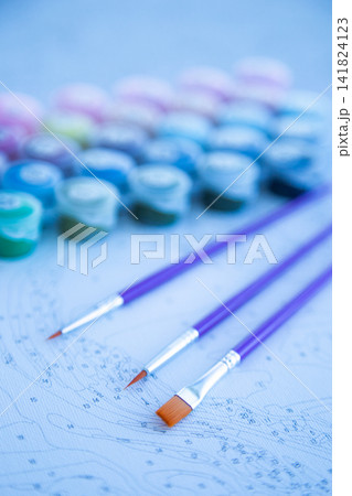 Three brushes with purple handles lie on a numbered canvas, and blurry paint jars are visible in the background. Concept of preparation for creativity, artistic tools, focus on the process. High 141824123