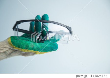 A hand wearing a latex work glove holds transparent safety glasses on a light background. Concept, vision care, workplace safety, demonstration of personal protective equipment. High quality photo A hand wearing a latex work glove holds transparent safety glasses on a light background. Concept, vision care, workplace safety, demonstration of personal protective equipment. High quality photo 141824132