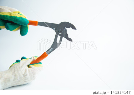 Two hands wearing protective gloves (green and yellow) hold adjustable pincers with bright orange handles horizontally on a white background. The concept of teamwork in renovation. High quality photo Two hands wearing protective gloves (green and yellow) hold adjustable pincers with bright orange handles horizontally on a white background. The concept of teamwork in renovation. High quality photo 141824147