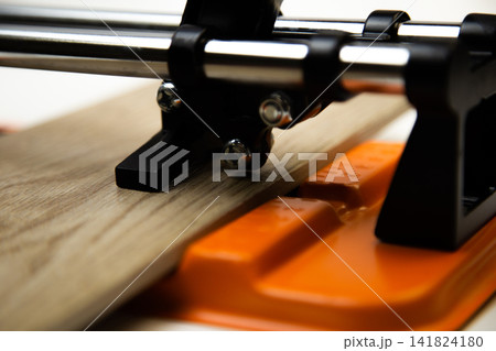 Close-up of an orange mechanical tile cutter, which fixed ceramic tiles of wooden colors, above it is a cutting disc on a white background. The concept of renovation, tiling, construction work. High 141824180