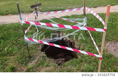 Deep dark hole in the ground cordoned with red and white caution tape on wooden stakes. 141824759
