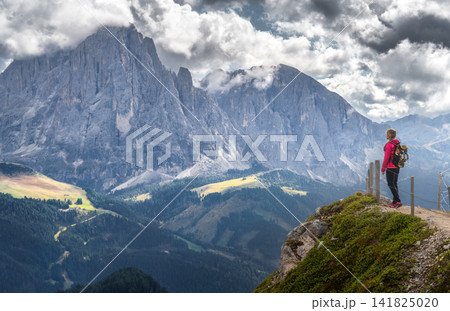 Woman with backpack standing on cliff edge looking at mountains 141825020
