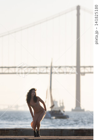 Woman poses by the water during sunset with a bridge in the background and a sailboat near the shore Woman poses by the water during sunset with a bridge in the background and a sailboat near the shore 141825180