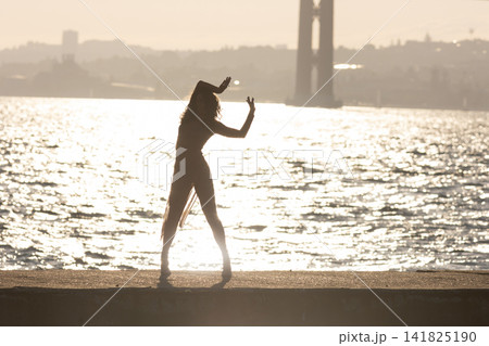 Dancer performs by the water as the sun sets behind a bridge in the background 141825190