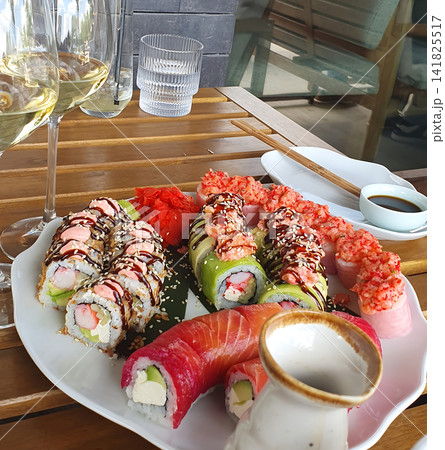 Delicious sushi set platter with various gourmet rolls, white wine glasses, and soy sauce served on a wooden terrace table Delicious sushi set platter with various gourmet rolls, white wine glasses, and soy sauce served on a wooden terrace table 141825517