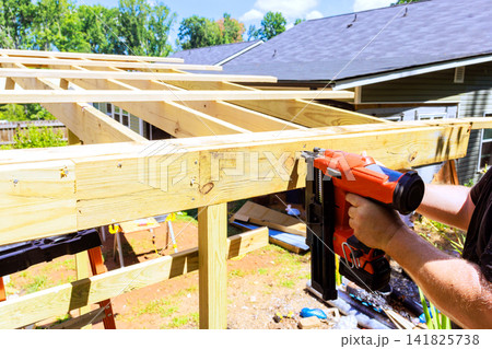 Handyman uses air nail gun to attach wooden beams while constructing frame structure in backyard Handyman uses air nail gun to attach wooden beams while constructing frame structure in backyard 141825738