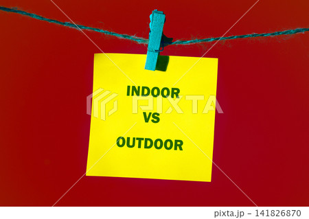 A yellow sticky note is clipped to a string hanging against a red background. The words indoor vs outdoor are clearly written in black letters on the note A yellow sticky note is clipped to a string hanging against a red background. The words indoor vs outdoor are clearly written in black letters on the note 141826870