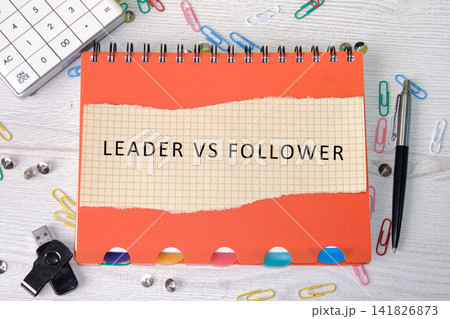 A notepad shows the words leader vs follower. Nearby are a pen, paper clips, a USB drive, and a calculator. This scene depicts an office setting and work thoughts 141826873