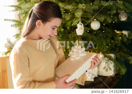 Teen girl holding Christmas gift beside decorated tree at home Teen girl holding Christmas gift beside decorated tree at home 141826917