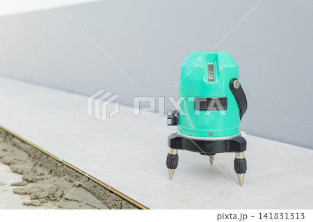 A green laser level on a tripod projects beams across a white and gray floor, ensuring precision while laying ceramic tiles in a construction setting A green laser level on a tripod projects beams across a white and gray floor, ensuring precision while laying ceramic tiles in a construction setting 141831313