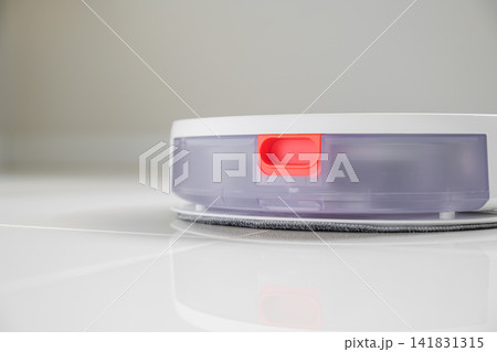 White automatic floor cleaner with side brush on tile flooring. Back view shows clear dustbin and red release button on white surface. Efficient smart home cleaning device for dust White automatic floor cleaner with side brush on tile flooring. Back view shows clear dustbin and red release button on white surface. Efficient smart home cleaning device for dust 141831315