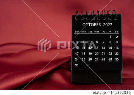 Black desk calendar for October 2027 on a red color satin or silk textile background. 141832030