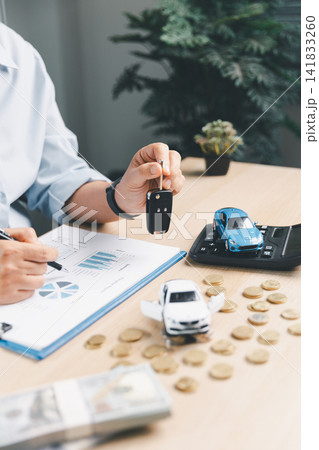 Business person holding car key while reviewing vehicle finance report with toy cars, calculator, coins, and cash, representing auto loan approval and car insurance. Business person holding car key while reviewing vehicle finance report with toy cars, calculator, coins, and cash, representing auto loan approval and car insurance. 141833260