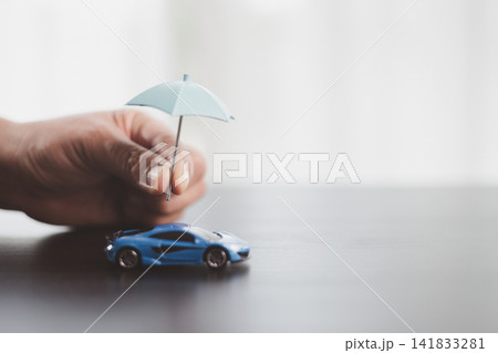 Hand holding a small umbrella above a blue model car symbolizes vehicle insurance, asset protection, accident coverage, risk management, ownership security, and responsible financial planning. Hand holding a small umbrella above a blue model car symbolizes vehicle insurance, asset protection, accident coverage, risk management, ownership security, and responsible financial planning. 141833281