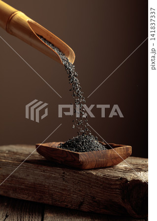 Grains of black sesame are poured with a wooden spoon in the wooden dish. Grains of black sesame are poured with a wooden spoon in the wooden dish. 141837337