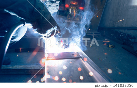 Welder welding metal with argon arc welding machine and has welding sparks and smoke. Man wear protective gloves. Safety in industrial workplace. Welder working with safety. Steel industry technology. 141838350