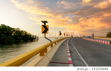 Curved coastal bridge with solar powered warning signs and streetlights at golden sunset, featuring modern transportation infrastructure, scenic seascape, mangrove shoreline and dramatic evening sky. Curved coastal bridge with solar powered warning signs and streetlights at golden sunset, featuring modern transportation infrastructure, scenic seascape, mangrove shoreline and dramatic evening sky. 141839088