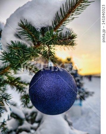 The Christmas tree is covered in snow and has a shiny ornament hanging from a branch. The sun sets in the background, marking the holiday season 141840323