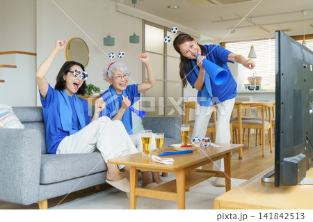 リビングのソファに並んで座ってテレビでサッカー日本代表を応援するミドル世代の日本人女性3人 リビングのソファに並んで座ってテレビでサッカー日本代表を応援するミドル世代の日本人女性3人 141842513