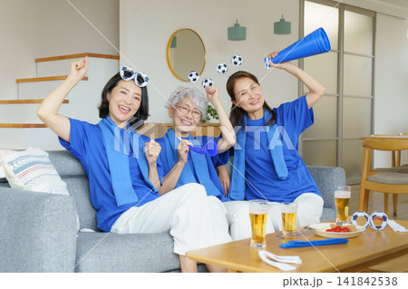 リビングのソファに並んで座ってテレビでサッカー日本代表を応援するミドル世代の日本人女性3人 141842538