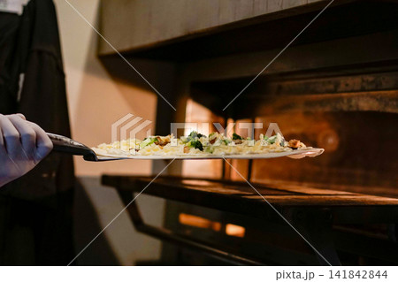 A fresh pizza on a metal peel being placed into a heated commercial oven. Professional restaurant kitchen atmosphere showcasing real culinary preparation. A fresh pizza on a metal peel being placed into a heated commercial oven. Professional restaurant kitchen atmosphere showcasing real culinary preparation. 141842844