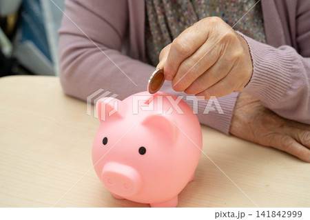 Retired elderly woman putting coins money in piggy bank and worry about monthly expenses and treatment fee payment. 141842999