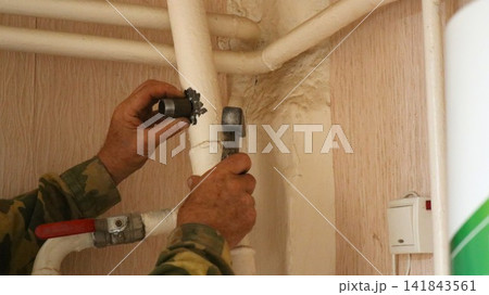 A worker adjusts iron heating pipes with a hammer. A light switch is visible on the wall. During the renovation, a plumber, wearing gloves, installs a new steel pipe. A worker adjusts iron heating pipes with a hammer. A light switch is visible on the wall. During the renovation, a plumber, wearing gloves, installs a new steel pipe. 141843561