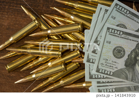 Scattered rifle cartridges beside hundred dollar bills on wooden surface Scattered rifle cartridges beside hundred dollar bills on wooden surface 141843910
