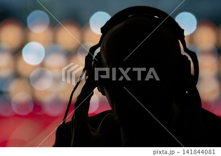 The dark silhouette of a man in headphones on the background of multicolored glare and lights of the city. Concept: government surveillance of people, secret special agent eavesdropping on 141844081