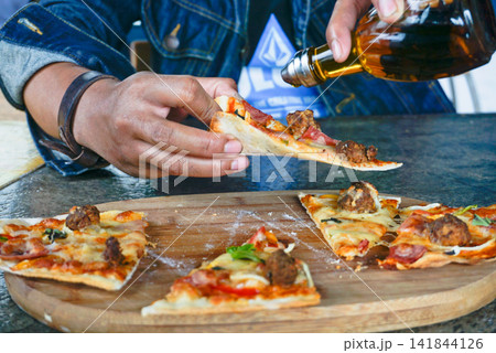 Close up of hands pouring chili oil onto pizza slice. Spicy topping, melted cheese, casual dining and lifestyle eating concept. 141844126