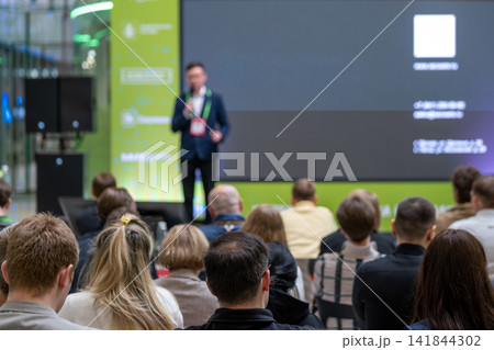 Audience watches business speaker beside large digital screen Audience watches business speaker beside large digital screen 141844302