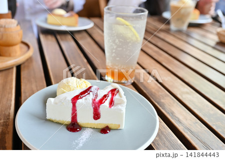 屋外カフェのウッドテーブルで楽しむ、ベリーソースのチーズケーキと爽やかなドリンク 141844443