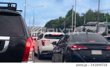 Rear view of multiple cars waiting in a heavy traffic jam on an urban highway. Highlighting red tail lights during a busy daytime commute. Background of green forest under blue sky and white clouds. 141844728