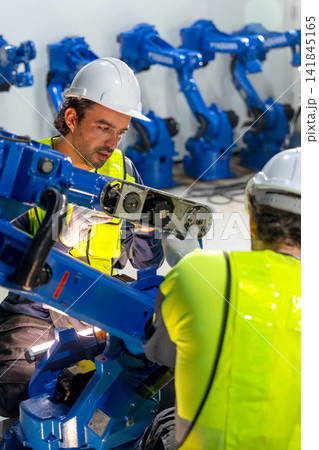 Two technicians in safety gear inspect blue robotic arm's internal components in industrial workshop with focused expressions Two technicians in safety gear inspect blue robotic arm's internal components in industrial workshop with focused expressions 141845165