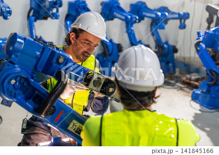 Two technicians in safety gear inspect blue robotic arm's internal components in industrial workshop with focused expressions 141845166