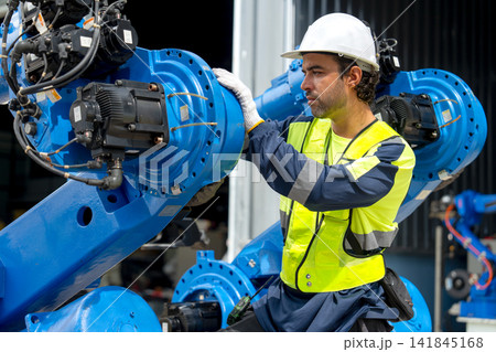 Male technician in safety gear inspects blue robotic arm with focused expression in industrial workshop 141845168