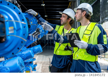 Two technicians in high-visibility vests examine blue industrial machinery with focused expressions 141845172