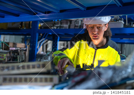 Female technician in bright yellow safety jacket examines mechanical part under blue industrial shelving Female technician in bright yellow safety jacket examines mechanical part under blue industrial shelving 141845181