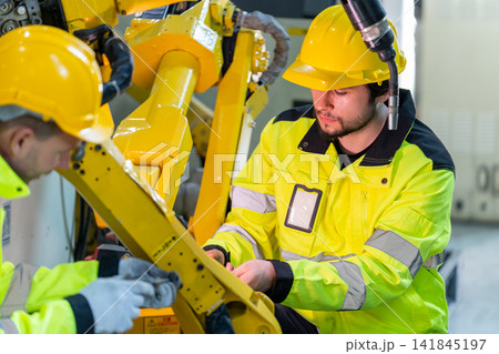 Two technicians in yellow hard hats and safety jackets adjust large robotic arm joint in industrial workshop with visible cables and tools Two technicians in yellow hard hats and safety jackets adjust large robotic arm joint in industrial workshop with visible cables and tools 141845197
