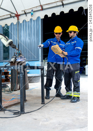 Two engineers in blue uniforms and yellow hard hats operate robotic welding arm in industrial workshop Two engineers in blue uniforms and yellow hard hats operate robotic welding arm in industrial workshop 141845208