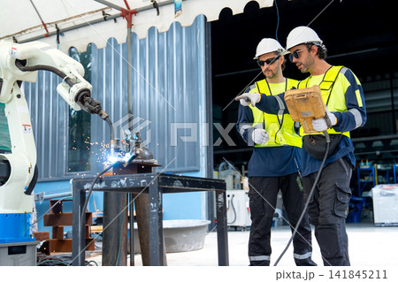Two engineers in safety gear operate robotic welding arm with sparks flying in industrial workshop 141845211