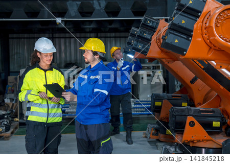 Three technicians in hard hats inspect orange robotic arm's internal components in industrial workshop with focused expressions Three technicians in hard hats inspect orange robotic arm's internal components in industrial workshop with focused expressions 141845218
