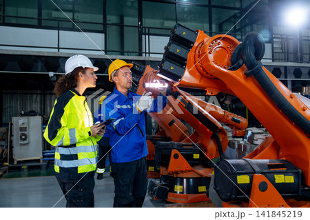 Two technicians in hard hats inspect orange robotic arm's internal components with focused expressions in modern factory Two technicians in hard hats inspect orange robotic arm's internal components with focused expressions in modern factory 141845219