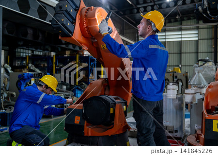 Two technicians in blue uniforms and yellow hard hats inspect orange robotic arm's joint with focused expressions in industrial workshop 141845226