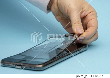 Female hands peel off the broken protective glass of a smartphone on a blue background, close-up, selective focus. Concept: glass replacement, phone repair with your own hands. Female hands peel off the broken protective glass of a smartphone on a blue background, close-up, selective focus. Concept: glass replacement, phone repair with your own hands. 141845498
