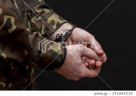 A military man in camouflage is handcuffed on a dark background. Concept: war criminal, prisoner of war, tribunal for deserters, traitor to the motherland. A military man in camouflage is handcuffed on a dark background. Concept: war criminal, prisoner of war, tribunal for deserters, traitor to the motherland. 141845518