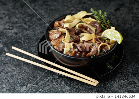 Shimeji and shiitake mushrooms stir-fried with soy sauce, garlic and thyme, served with noodles and lime slice in a black ceramic bowl. 141846634