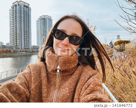 Woman wearing a cozy brown coat and sunglasses taking a selfie beside an urban waterfront in autumn, with modern high-rise buildings and ornamental grasses. Contemporary city lifestyle, fall fashion. 141846772
